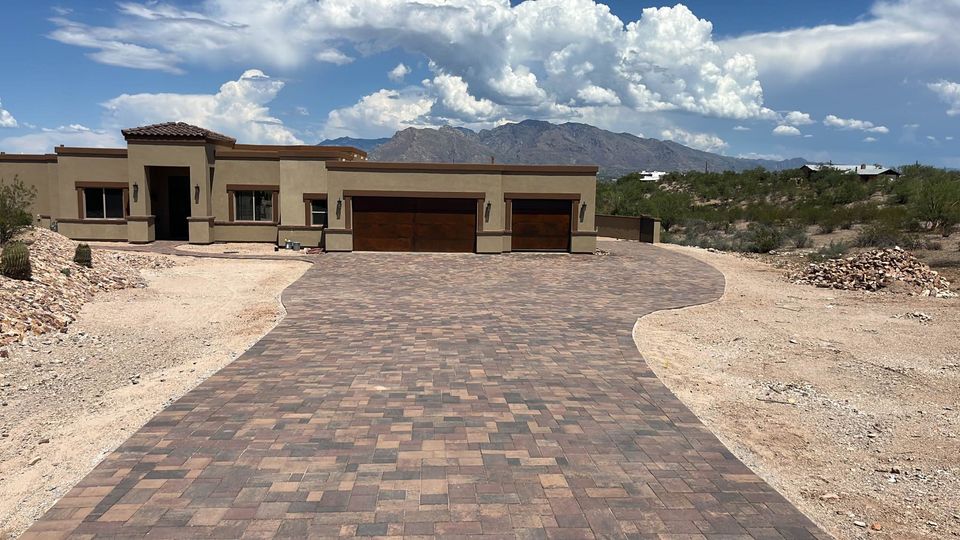 A modern, single-story house with tan walls and a large, paved driveway—showcasing expert paver installation. The home sits amid a desert landscape and mountains under a partly cloudy sky near Phoenix.