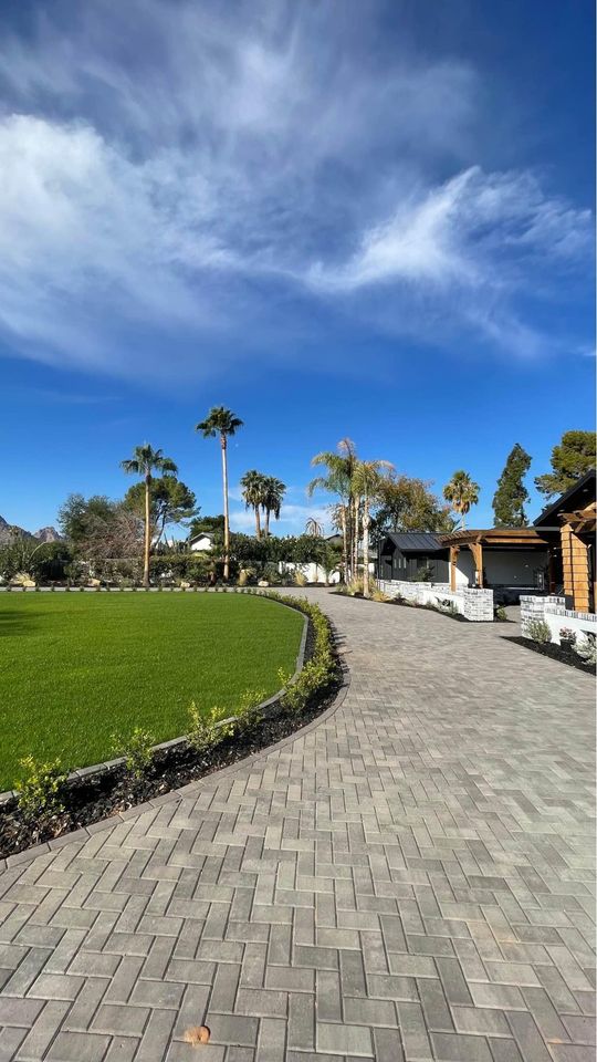 A wide stone walkway, the result of expert paver installation Phoenix, curves alongside a neatly trimmed green lawn with palm trees and shrubs. Buildings with wooden and white accents are visible to the right under a bright blue sky with wispy clouds.