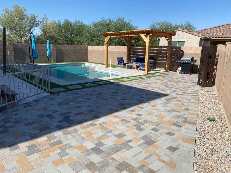 A backyard features a paved patio by Paver Installation Phoenix, a swimming pool with a safety fence, a wooden pergola with outdoor seating, barbecue grill, blue umbrellas, and desert landscaping with stone and trees in the background.