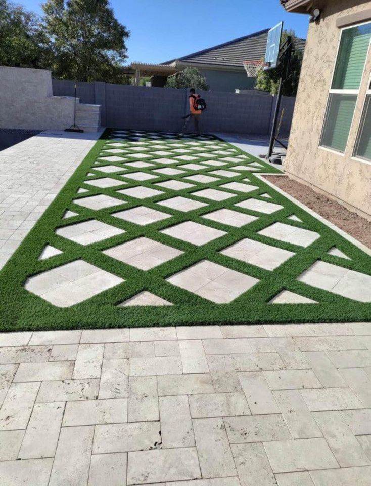 A backyard patio features a geometric pattern of green artificial grass and light stone tiles, arranged in a crisscross design—a great example of expert paver installation Phoenix homeowners appreciate. A person uses a leaf blower; a basketball hoop is in the background.