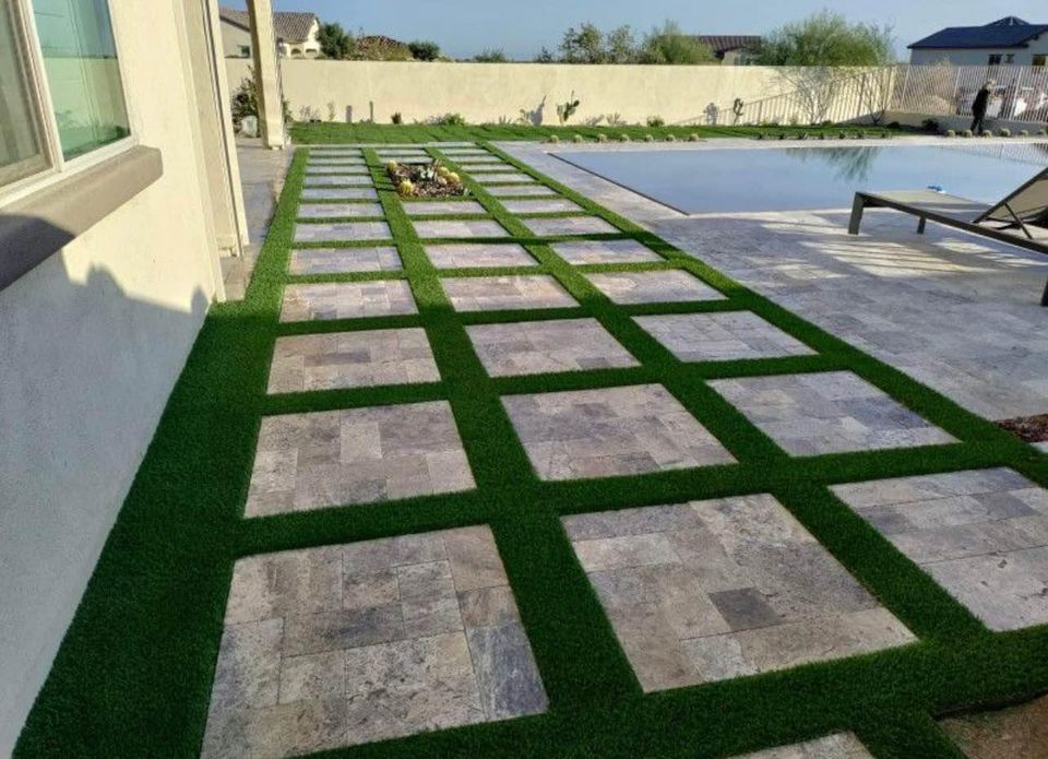 A modern patio with square stone tiles arranged in a grid pattern, separated by green grass strips, showcases expert paver installation Phoenix style—set beside a swimming pool and lounge chairs in a stylish backyard.