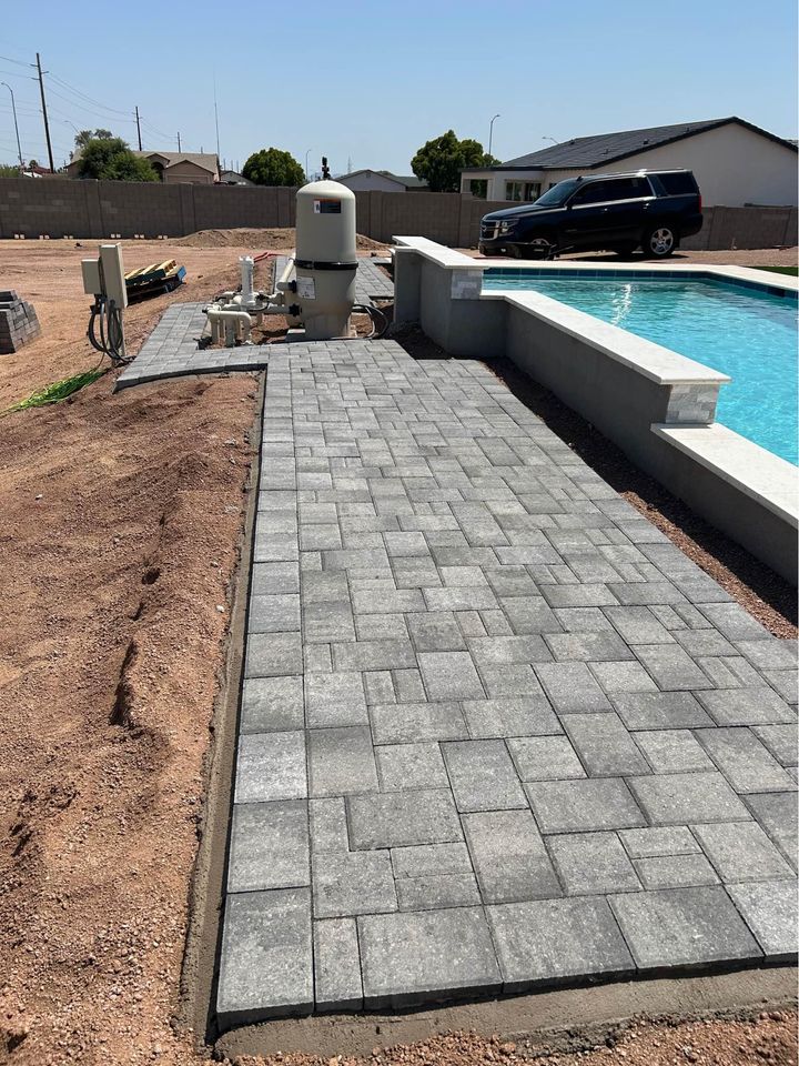 A newly paved gray stone walkway, showcasing expert paver installation Phoenix, runs beside a backyard swimming pool bordered by unfinished landscaping and pool equipment. A black SUV is parked in the background near a house.