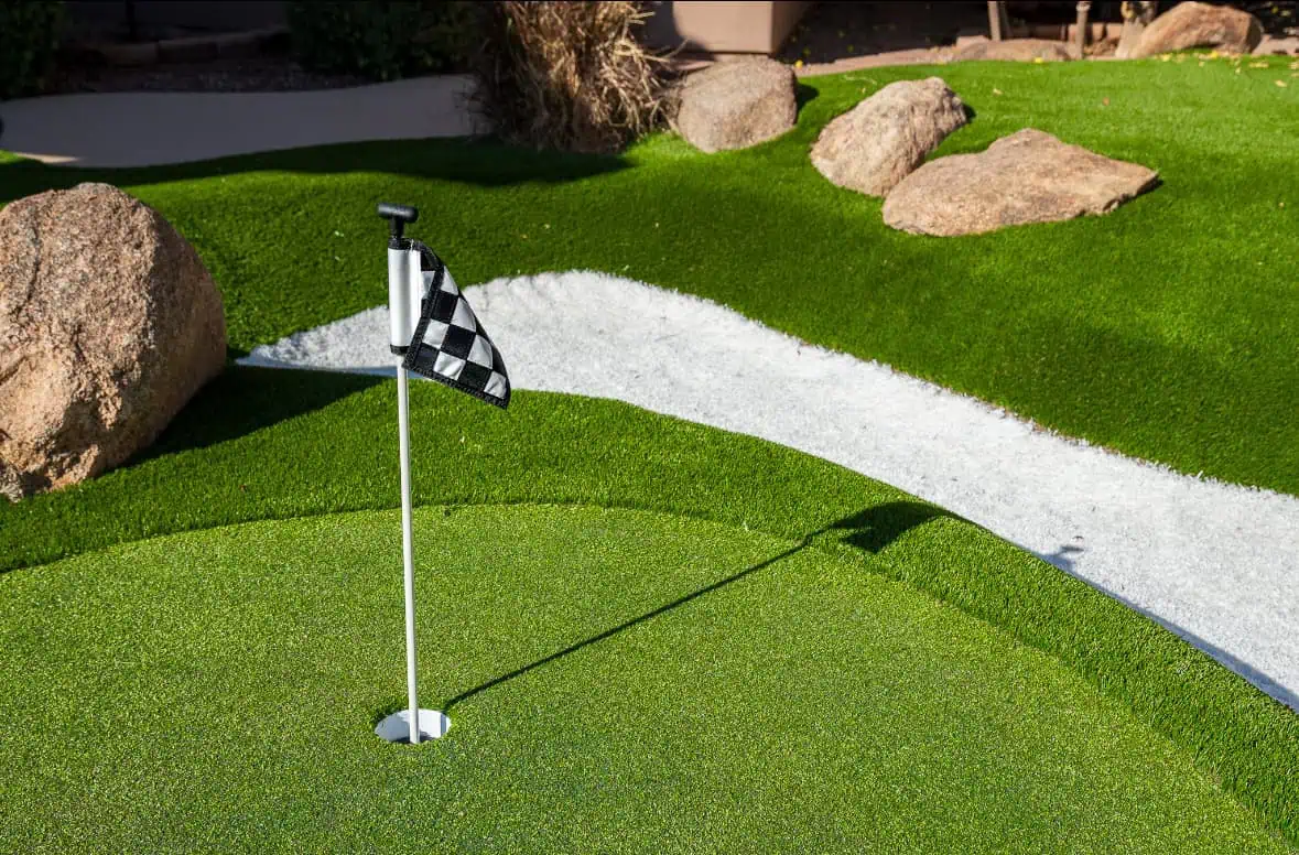 A close-up of a mini golf green with a black-and-white checkered flag in the hole. There are large rocks, artificial grass, and a white sand bunker in the background.