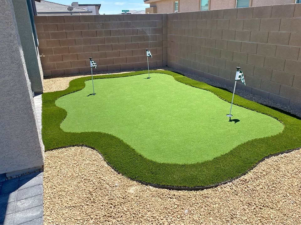 A small backyard putting green with three golf holes and flags, surrounded by artificial turf and gravel, enclosed by tan brick walls—expertly crafted by a top-rated artificial grass installer in Phoenix.