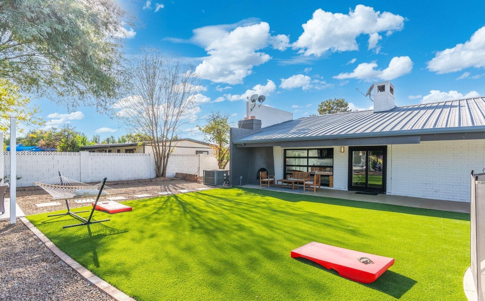 A backyard with lush lawn by a top-rated artificial grass installer Phoenix, red cornhole boards, a hammock, a covered patio with chairs and table, and a white brick fence under a blue sky with scattered clouds.