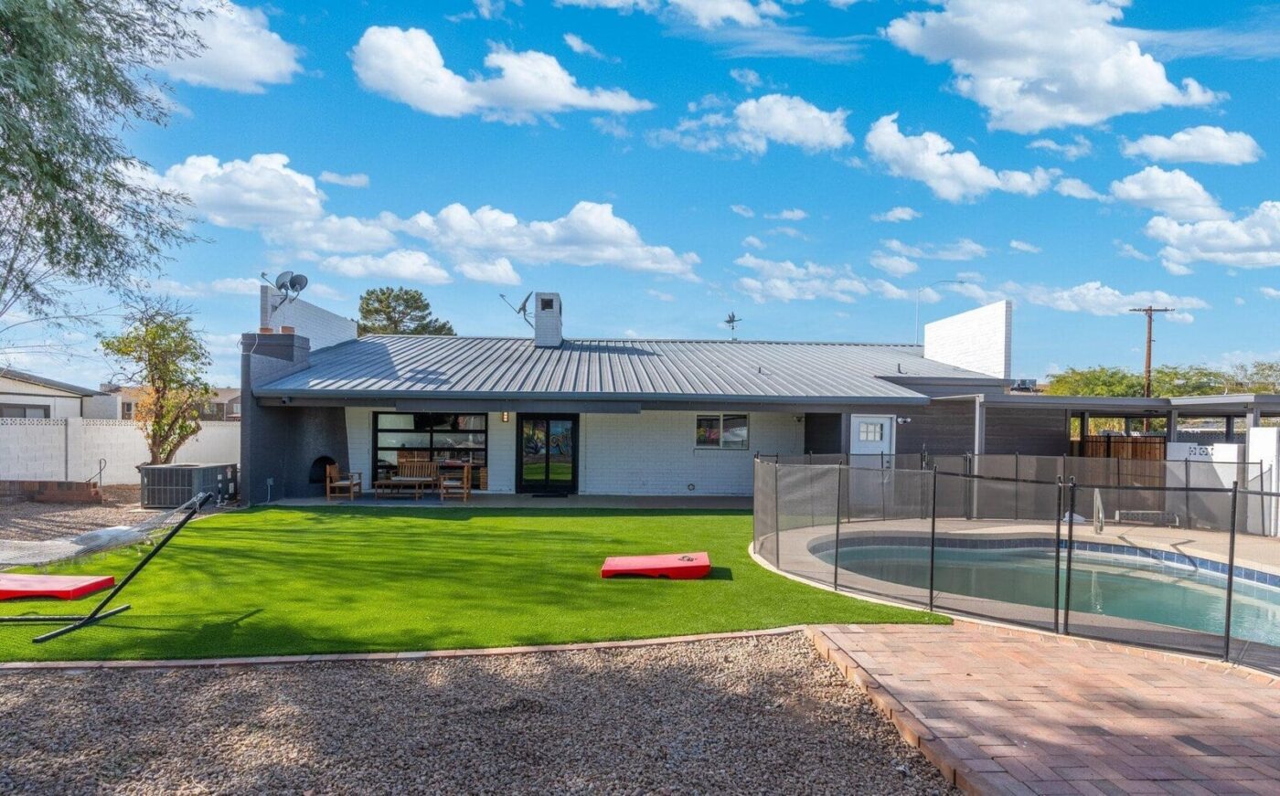 A backyard by a single-story house features top-rated artificial grass installed by a Phoenix expert, with a fenced oval swimming pool, patio furniture, hammock, and two red cornhole boards under a blue sky with scattered clouds.