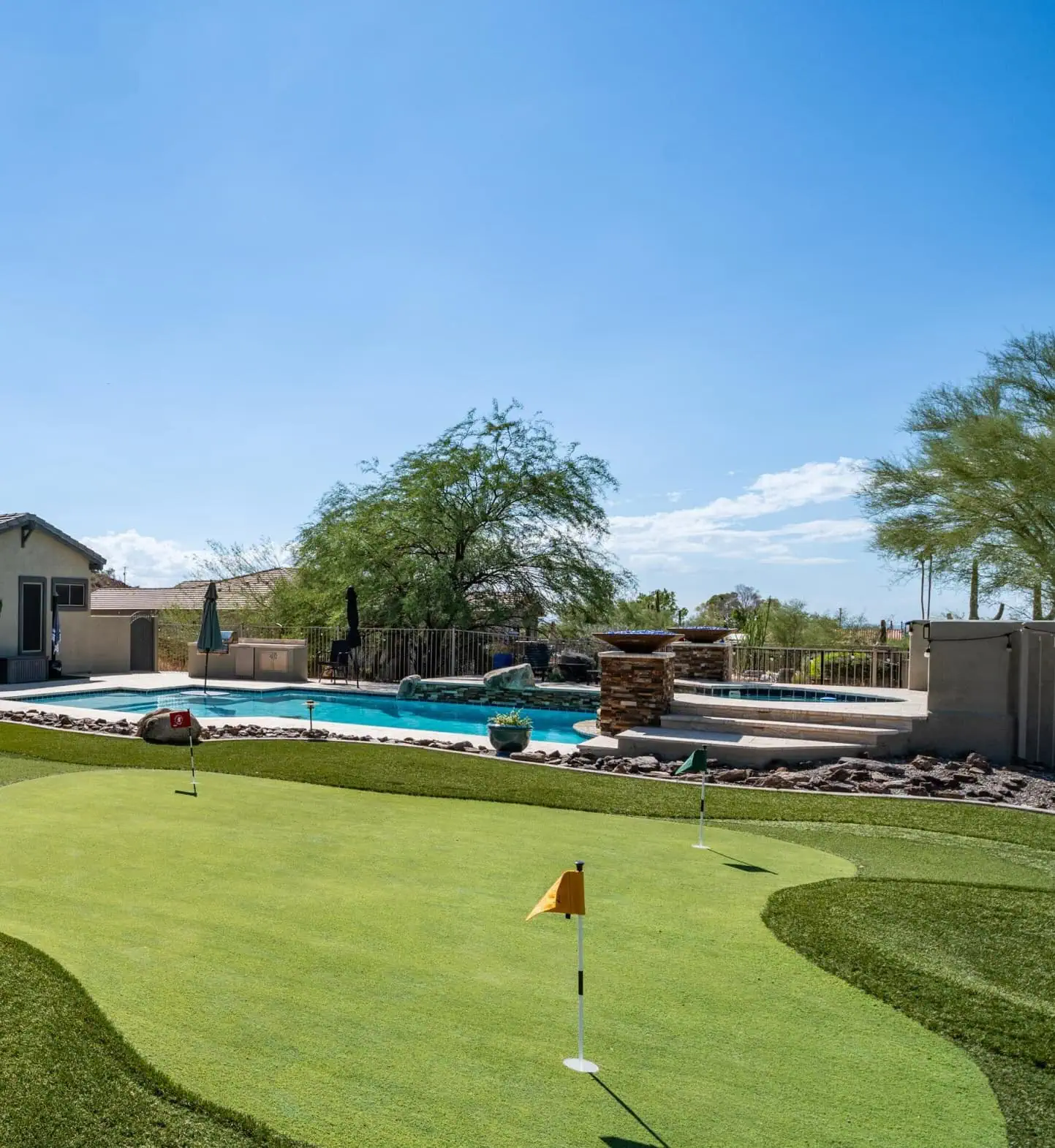 A backyard features a small putting green by a Top-Rated Artificial Grass Installer Phoenix, a swimming pool with clear water, lounge chairs, a stone waterfall feature, and desert landscaping under a bright blue sky.