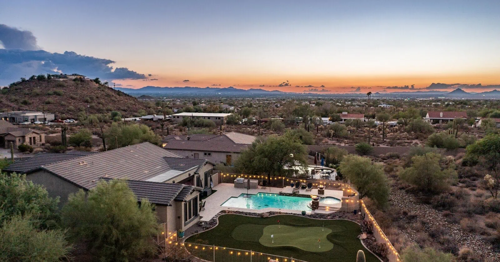 Aerial view of a modern house with a backyard pool, hot tub, and putting green by Top-Rated Artificial Grass Installer Phoenix, surrounded by desert landscaping and hills, at sunset with string lights and distant mountains.