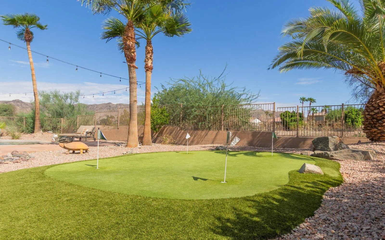 A backyard putting green with several golf flags by a top-rated artificial grass installer in Phoenix, surrounded by palm trees, desert landscaping, and a fence under a clear blue sky. String lights are hung between the trees.