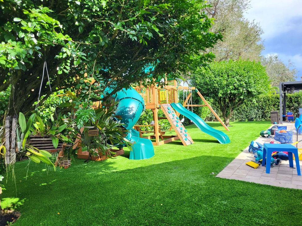 A backyard with Playground Artificial Grass Phoenix AZ, a blue and green playground set featuring slides and climbing walls under trees, plus a patio area with blue plastic chairs and scattered toys.