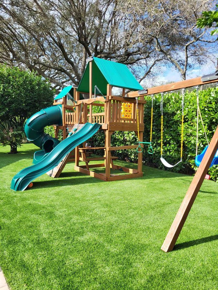 A wooden backyard playset with a green slide, green tube slide, swings, and climbing features sits on Playground Artificial Grass in Phoenix, AZ, surrounded by lush hedges and tall trees under a bright blue sky.