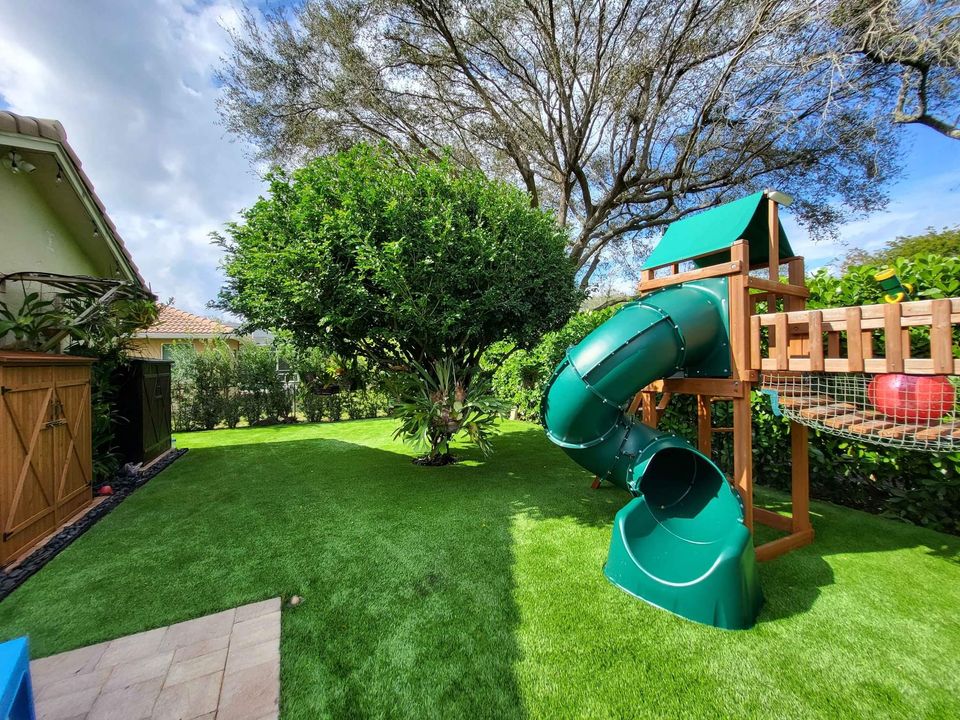A backyard with bright green playground artificial grass in Phoenix, AZ, a large leafy tree, and a wooden playset featuring a covered platform and a green spiral slide, surrounded by lush plants and a wooden fence.