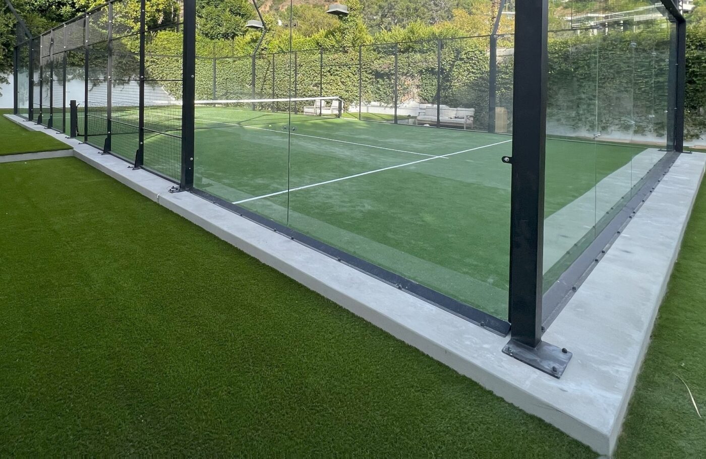 A modern outdoor padel court with green artificial grass installed by a top-rated artificial grass installer in Phoenix, is surrounded by clear glass walls and black metal posts on a sunny day with greenery and trees in the background.