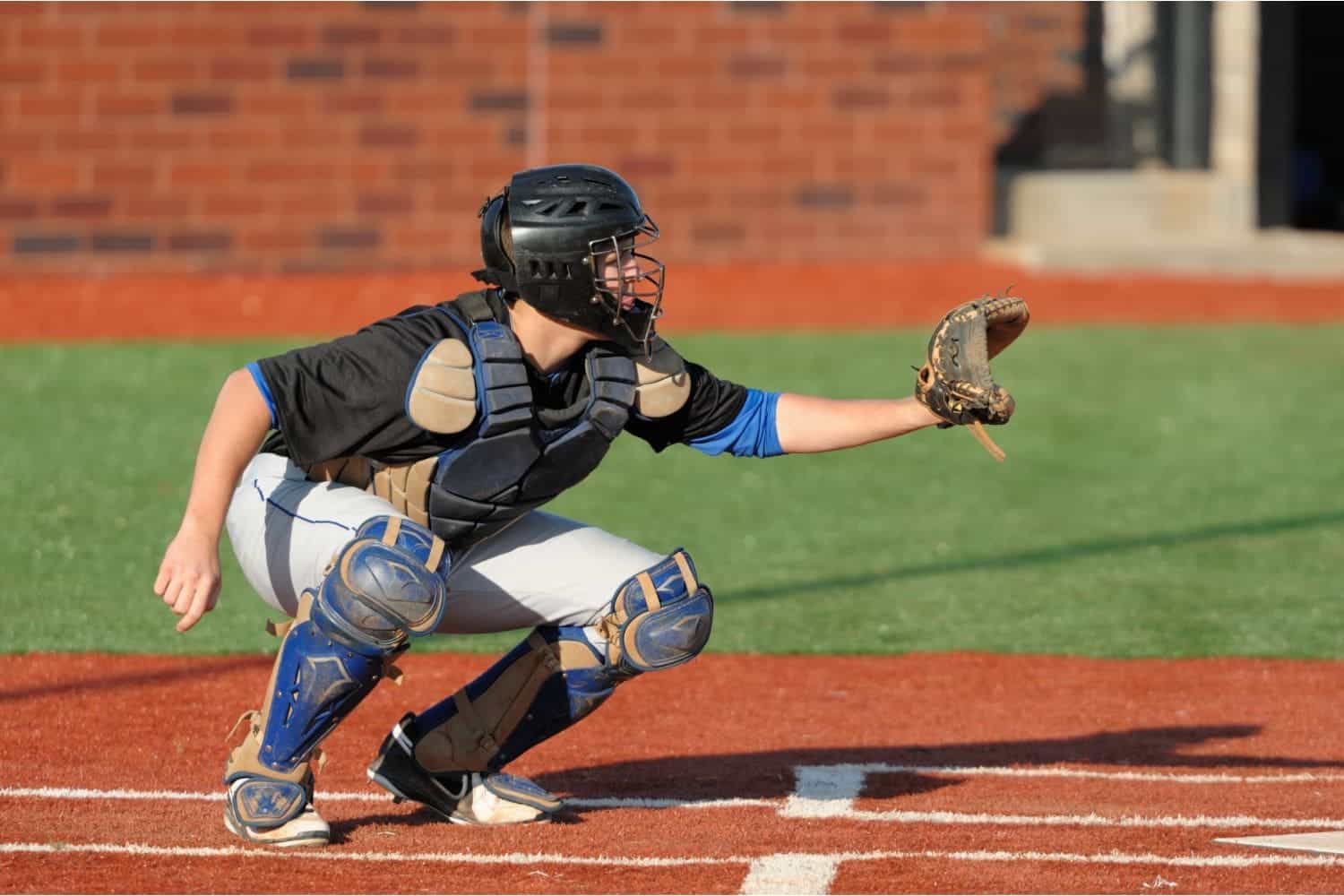 A baseball catcher in full gear crouches behind home plate with a mitt extended, ready to catch a pitch on a field installed by Top-Rated Artificial Grass Installer Phoenix, with a brick wall in the background.