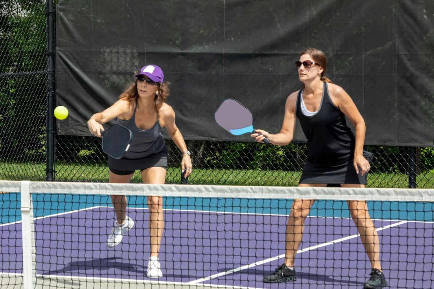 Two women play pickleball on an outdoor court featuring lush turf by a Top-Rated Artificial Grass Installer Phoenix; one is about to hit the ball with her paddle while the other stands ready. Both are wearing athletic clothing and sunglasses.