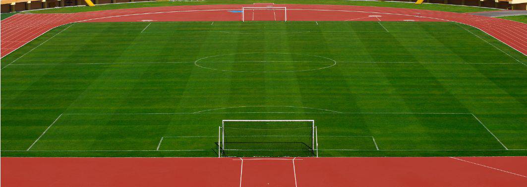 A view of an empty soccer field with lush green grass installed by a top-rated artificial grass installer Phoenix, white markings, and red running tracks. There are goals at both ends and stadium seating visible in the background.
