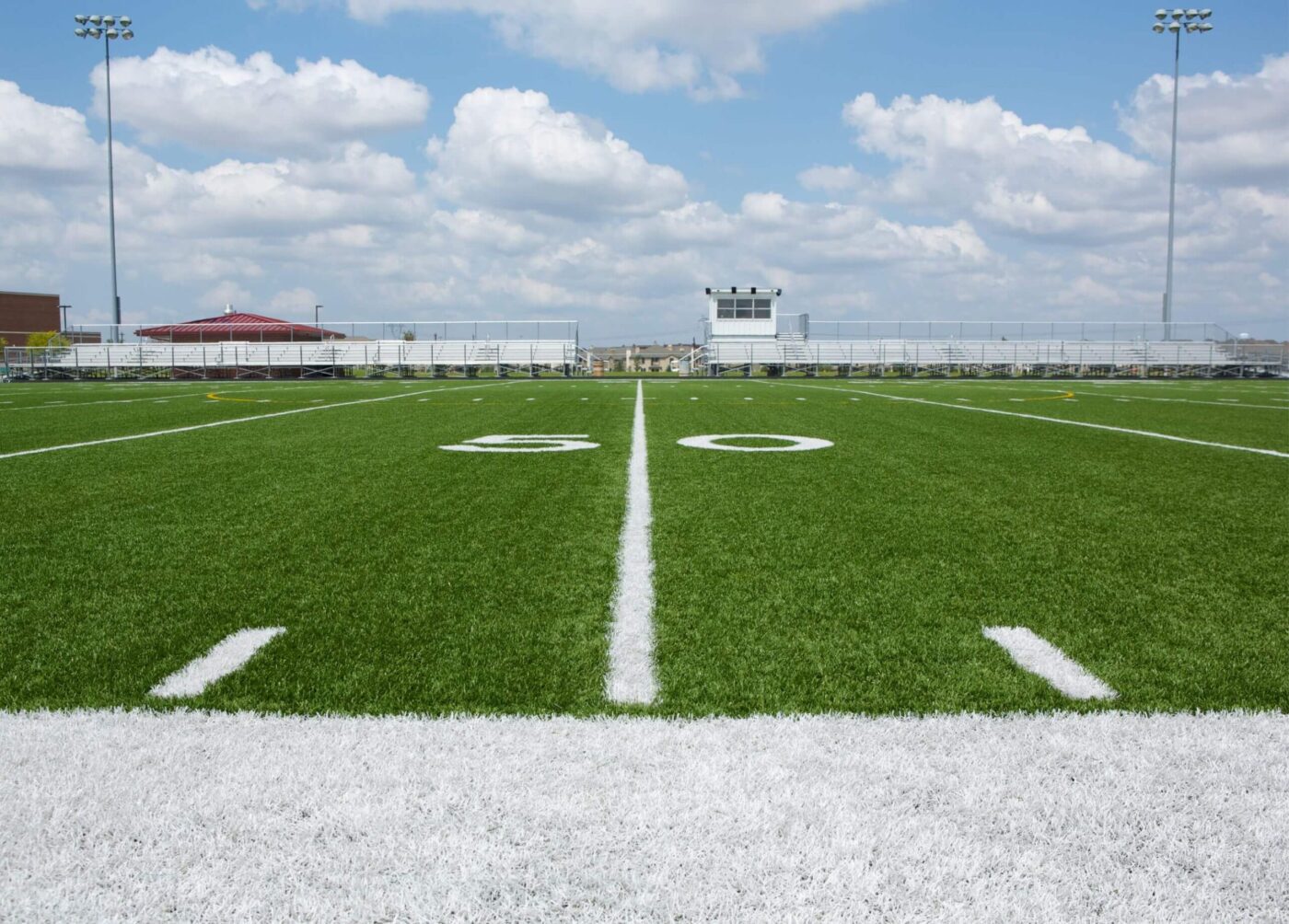 A football field seen from the 50-yard line, with bright green turf installed by a Top-Rated Artificial Grass Installer Phoenix, white markings, a scoreboard, and empty bleachers in the background under a partly cloudy sky.