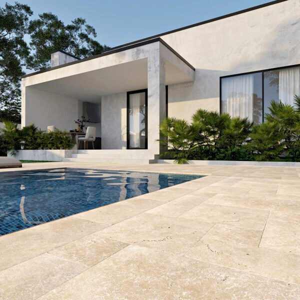 Modern house with large windows, a covered patio with outdoor furniture, and a swimming pool surrounded by beige stone tiles from expert Paver Installation Phoenix. Lush green plants line the patio’s edge beneath a clear, sunny sky.